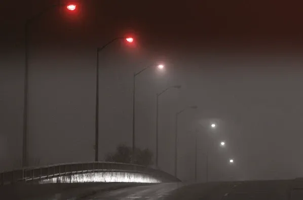 Strada deserta di notte avvolta nella nebbia, che sale su un ponte dal parapetto curvo, con una fila di lampioni accesi che si perde in lontananza
