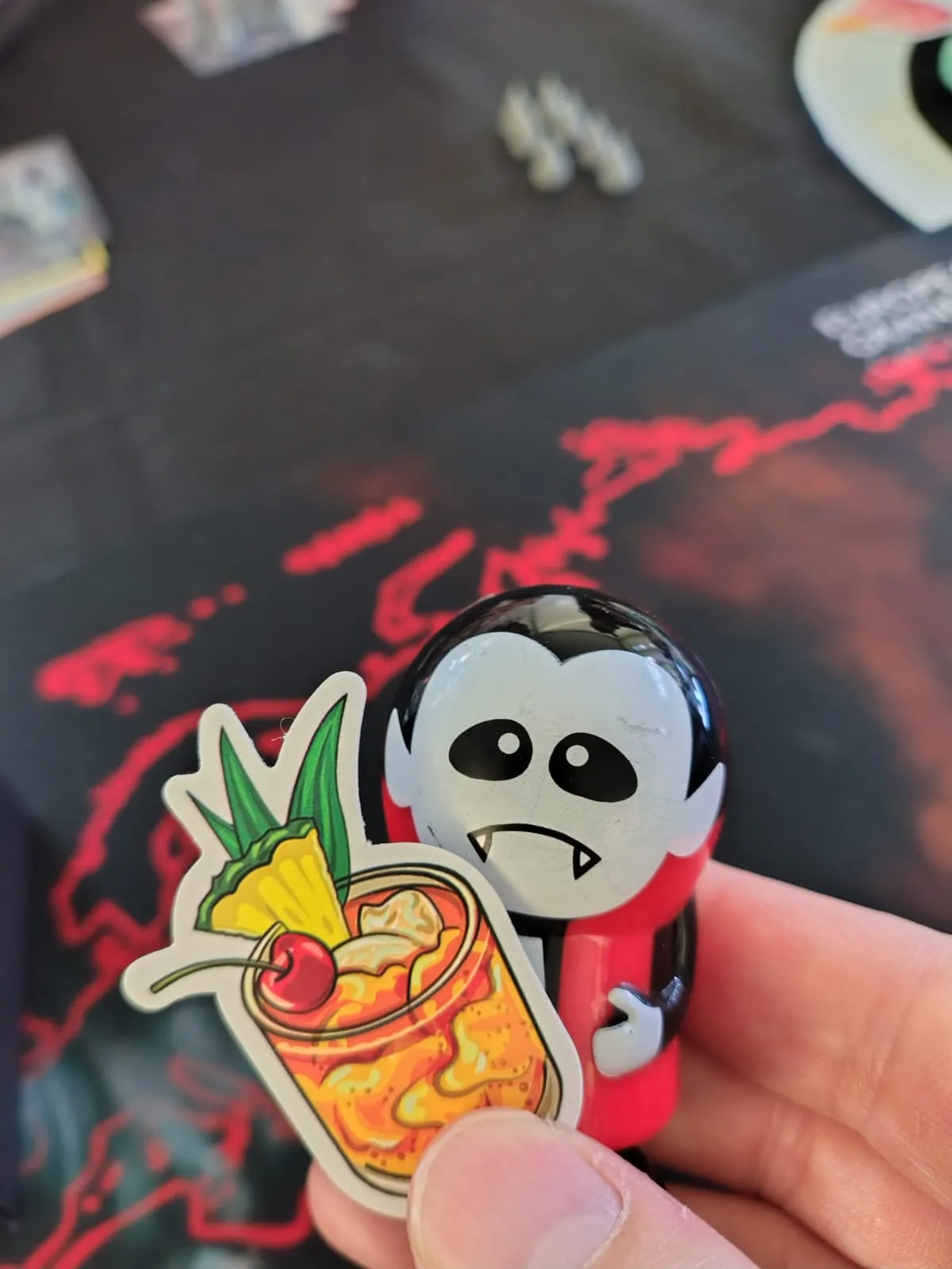 Un vampiretto giocattolo e un adesivo a forma di cocktail con ananas e ciliegia, tenuti in mano sopra il tappeto di gioco